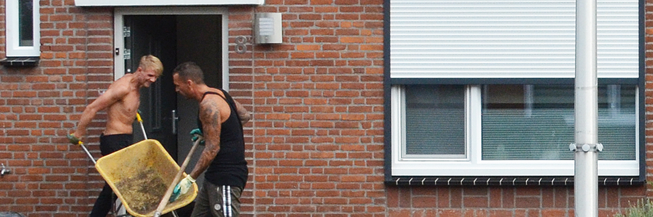 Eengezinswoning met bruine bakstenen muur, witte kozijnen en twee bouwvakker voor de voordeur met een kruiwagen