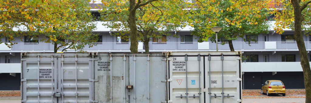 Appartementencomplex met daarvoor een boom, een container en een gele auto