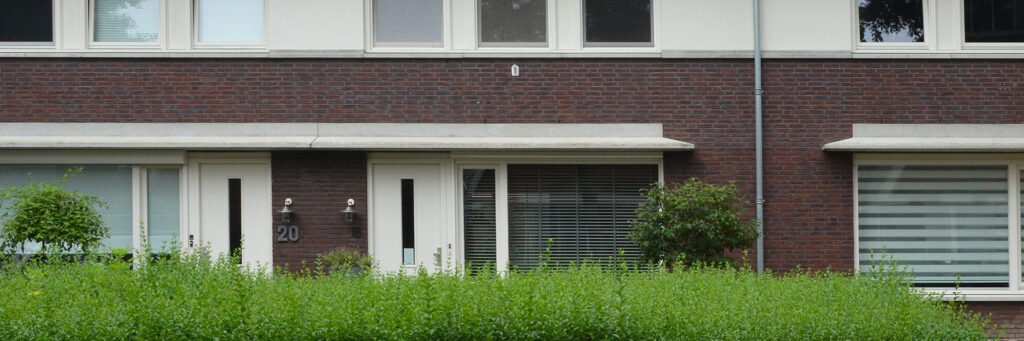 Eengezinswoning beneden verdieping, donkerbruine bakstenen muur en witte kozijnen en deur met een bosje ervoor