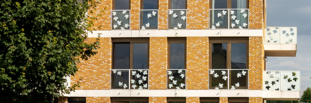 Appartementencomplex met gele bakstenen gevel, witte glazen balkons met blaadjes erop en een boom voor de deur