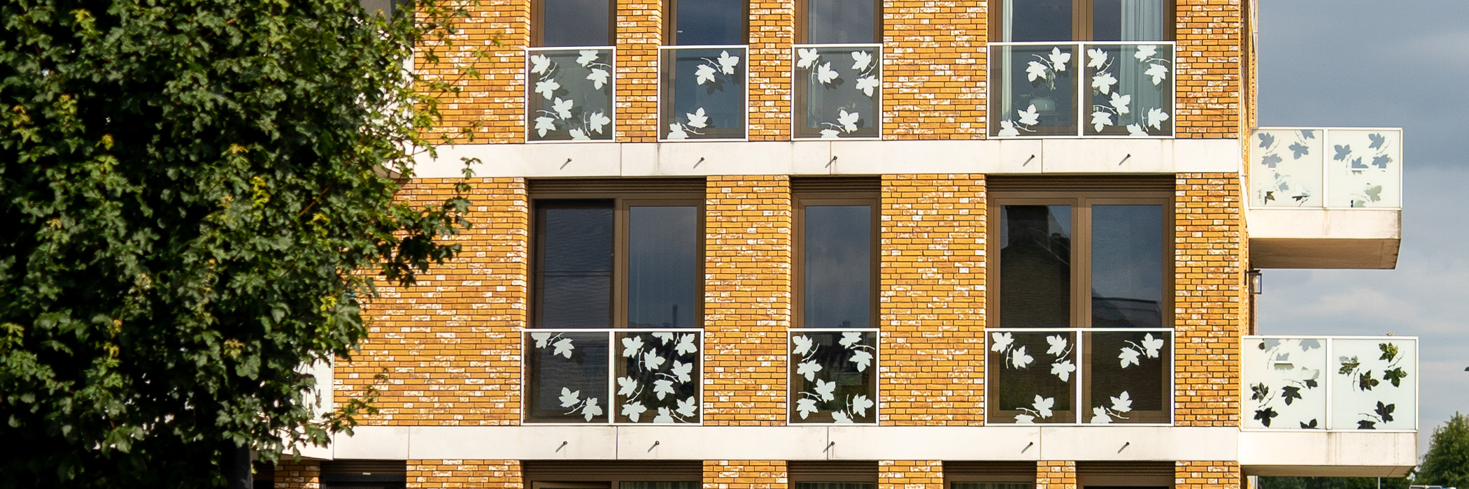 Appartementencomplex met gele bakstenen gevel, witte glazen balkons met blaadjes erop en een boom voor de deur