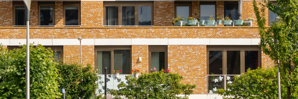 Woningen met bruin gele bakstenen gevel, bruine kozijnen en witte glazen balkons met blaadjes erop en bomen voor de deur