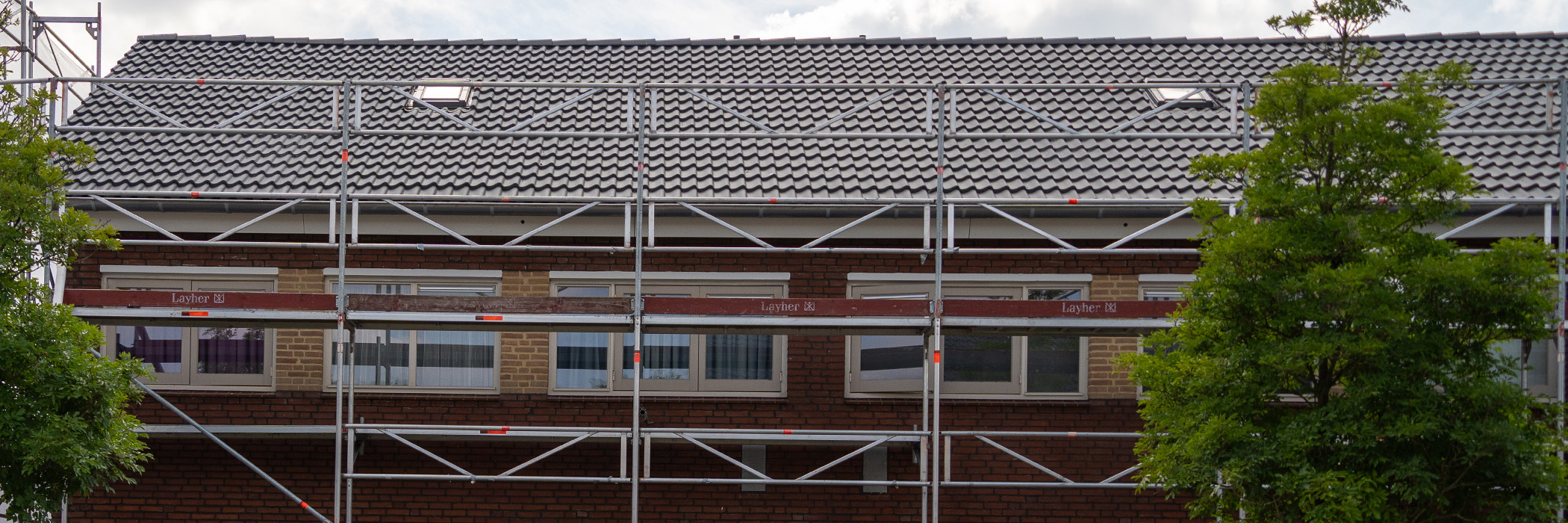 Woningen met bruin gele bakstenen gevel, crème kozijnen en een steiger ervoor met twee bomen
