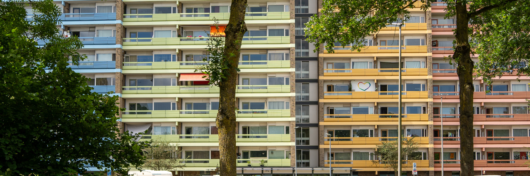 Appartemetencomplex met oranje, gele, groene en blauwe balkons en drie bomen voor de deur