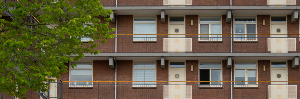 Appartementencomplex met bruine bakstenen gevel, crème ramen en deuren met een grijze gele balustrade met een boom ervoor