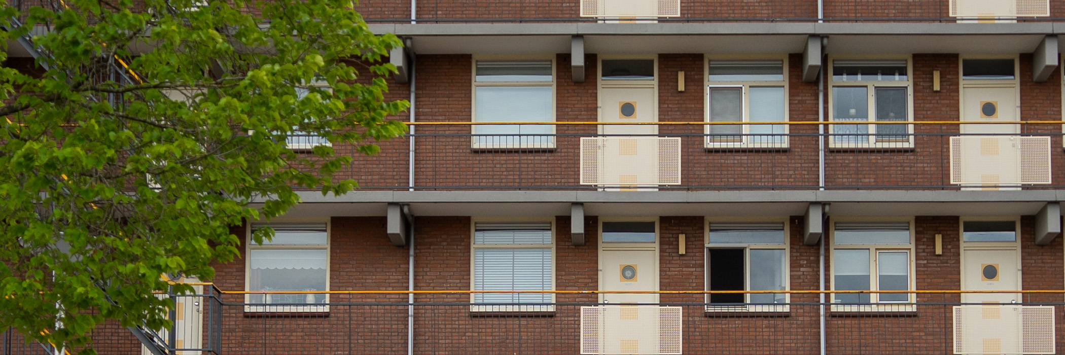 Appartementencomplex met bruine bakstenen gevel, crème ramen en deuren met een grijze gele balustrade met een boom ervoor