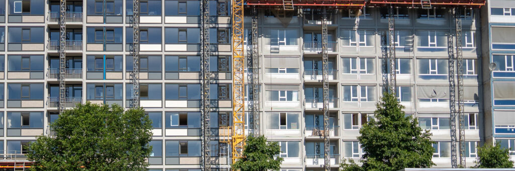 Appartementen met licht en donkergrijze gevel, witte en grijze kozijnen met blauwe vlakken met daarvoor een steiger en meerdere bomen
