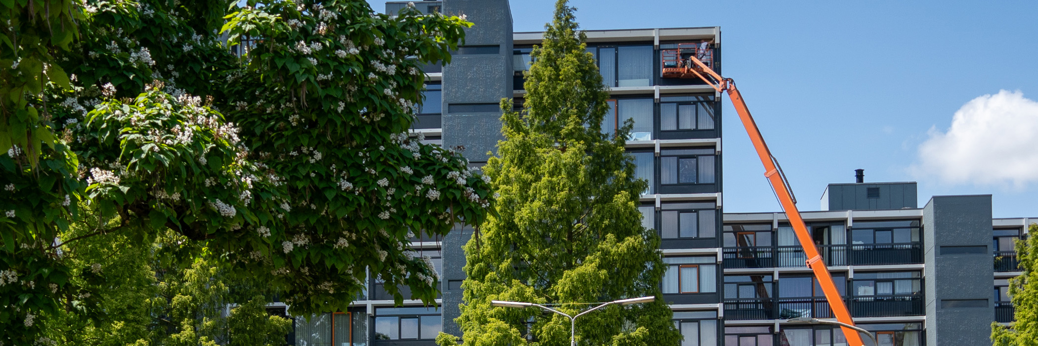 Appartementencomplex met grijze gevel, grijze kozijnen, een oranje hoogwerker ervoor die de ramen aan het schoonmaken is met bomen ervoor