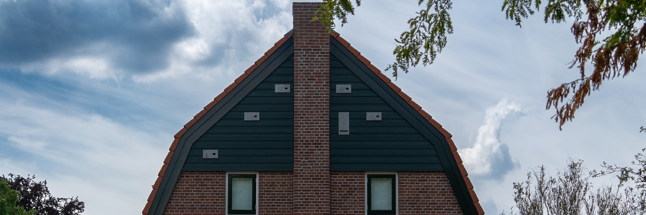 Woning waarvan de bovenste verdieping te zien is, met rode dakpannen, groene houten gevel in combinatie met bruine bakstenen en een schoorsteen met witte kozijnen en groene ramen