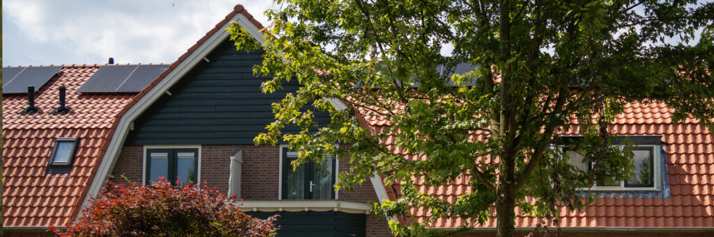 Woning waarvan de bovenste verdieping te zien is, met rode dakpannen, met zonnepanelen erop, groene houten gevel in combinatie met bruine bakstenen en een schoorsteen met witte kozijnen en groene ramen en bomen ervoor