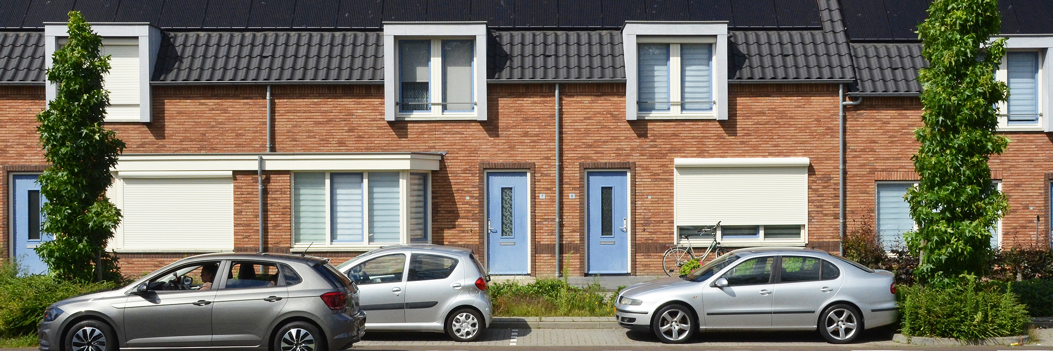 Woningen met bruine bakstenen gevel, witte kozijnen, blauwe deuren en auto's voor de deur met bomen