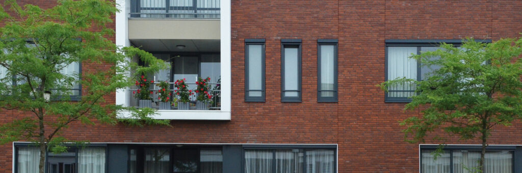 Appartementencomplex met donker rode bakstenen gevel, grijze kozijnen met witte balkons en twee bomen