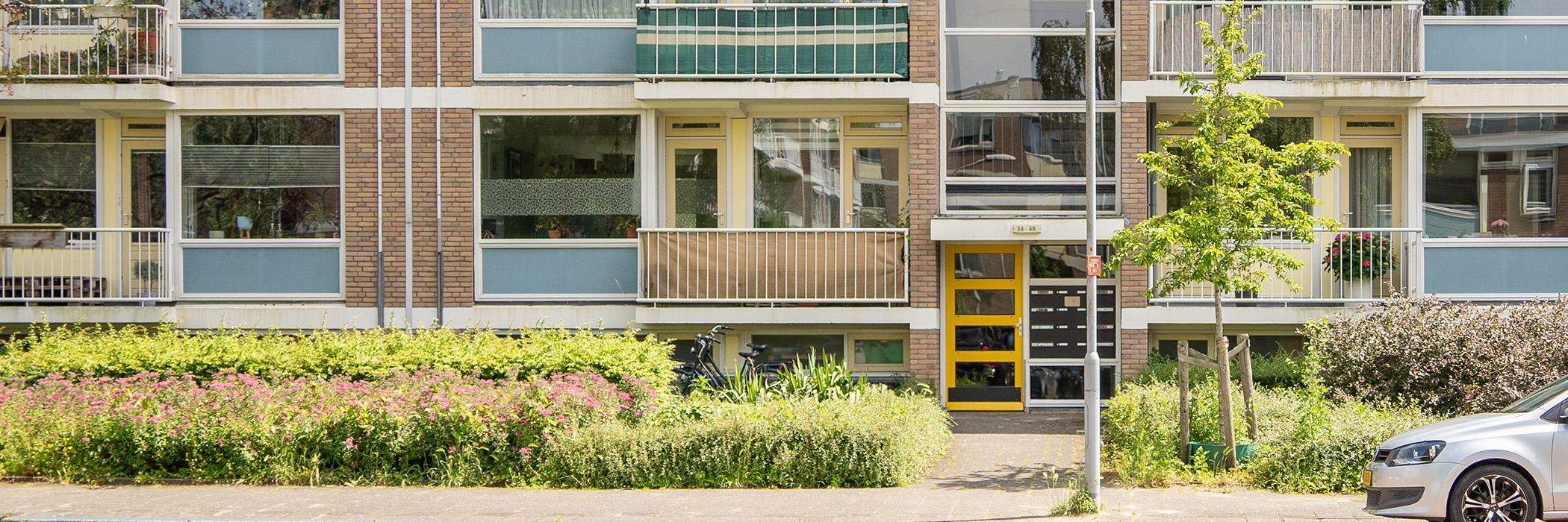 Appartementencomplex met bruine bakstenen gevel, witte kozijnen, met blauwe vlakken, gele deuren en ramen met een boom en een auto voor de deur