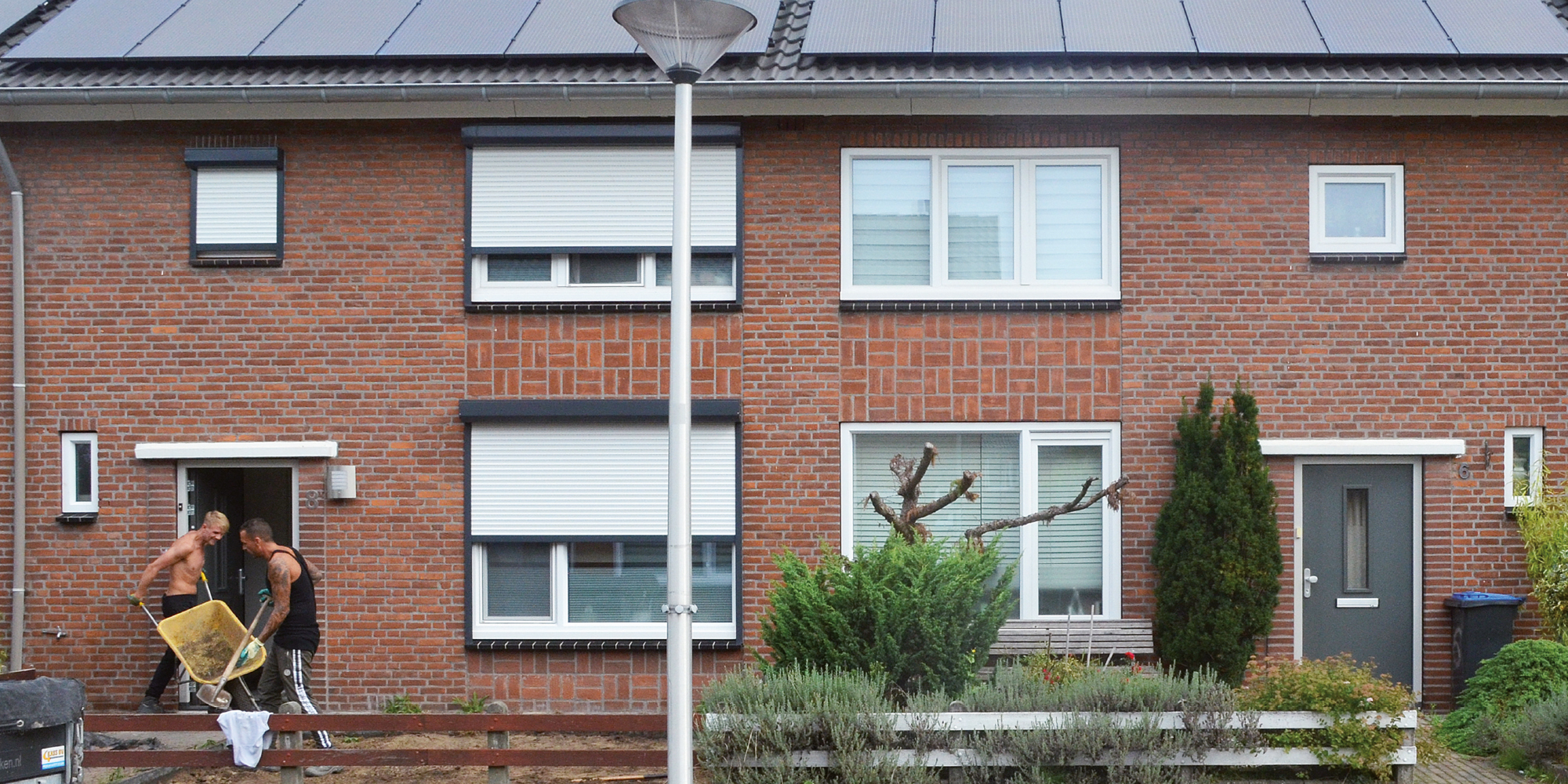 Woningen met bruine gevel, witte kozijnen, zonnepanelen op het dak, lantarenpaal en twee bouwvakkers in de deuropening