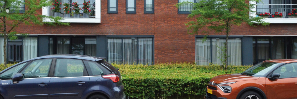 Woningen van bakstenen met een wit balkon op het balkon staan bloemen. Voor de woning staan groene bosjes.