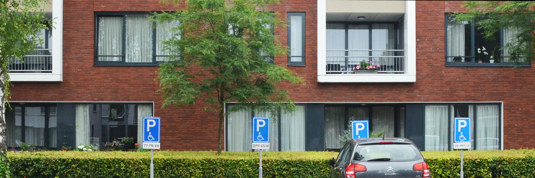 Appartementen met bruin/rode bakstenen gevel, grijze kozijnen en vier invalide parkeer borden voor de deur met bosjes waar een Citroen C3 voor staat