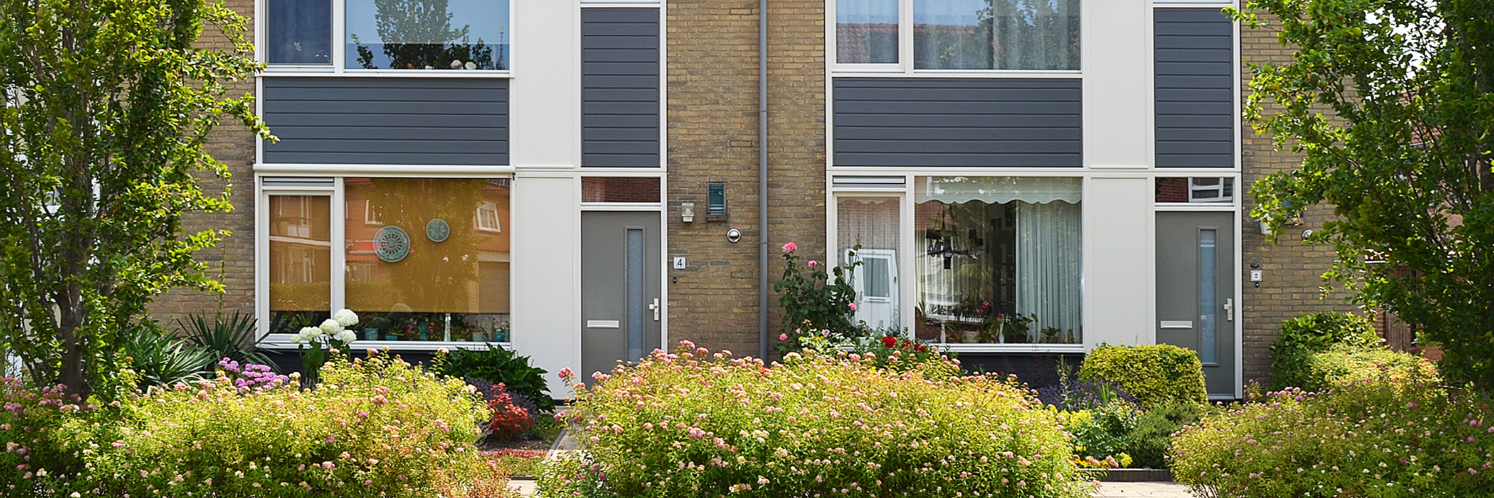 Woningen met bruine bakstenen gevel, witte kozijnen met grijze platen eronder, grijze deuren en planten voor de deur