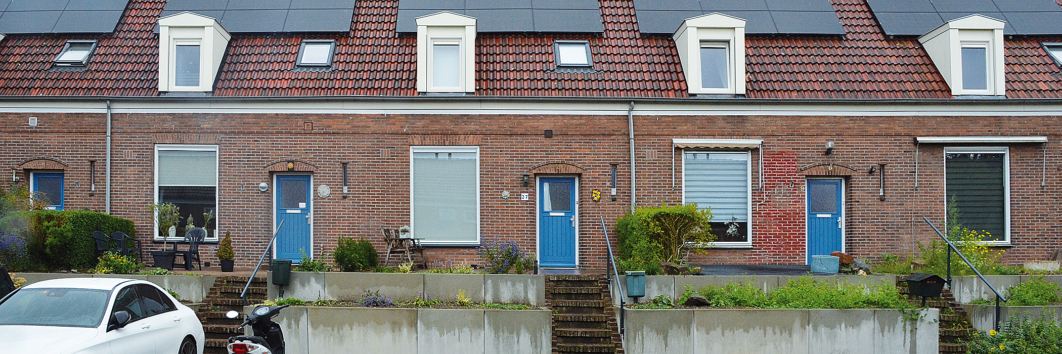 Woningen met bruine bakstenen gevel, rode dakpannen met zonnepanelen en dakramen met witte kozijnen en blauwe deuren met een witte auto en een scooter voor de deur