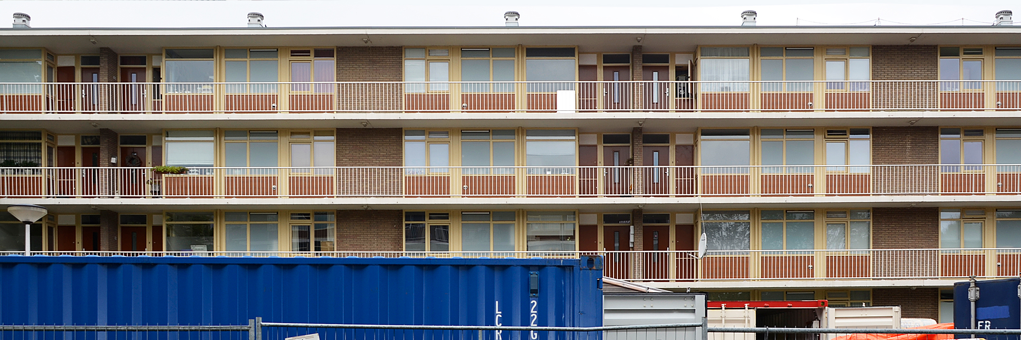 Appartementen met bruine bakstenen gevel, gele kozijnen en witte balustrade met containers voor de deur