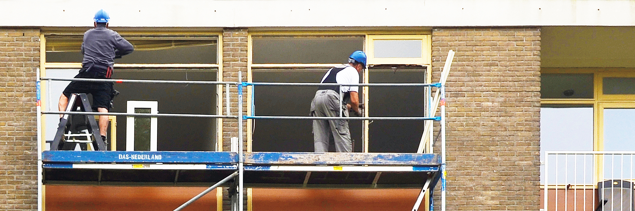Appartementen met bruine bakstenen gevel, met gele kozijnen en een steiger ervoor van das-nederland waar twee mannen op werken met een blauwe helm op