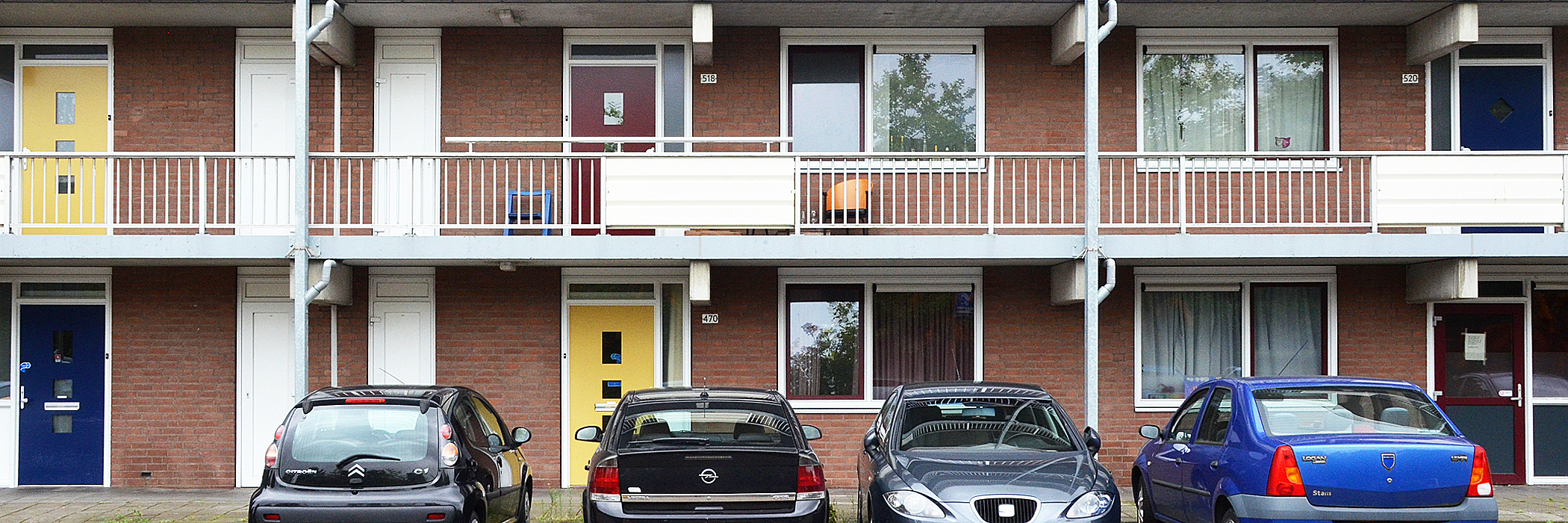 Appartementen met bruine bakstenen gevel, witte kozijnen, rode, gele en blauwe deuren met vier auto's voor de deur: Citroen C1, Opel, Seat