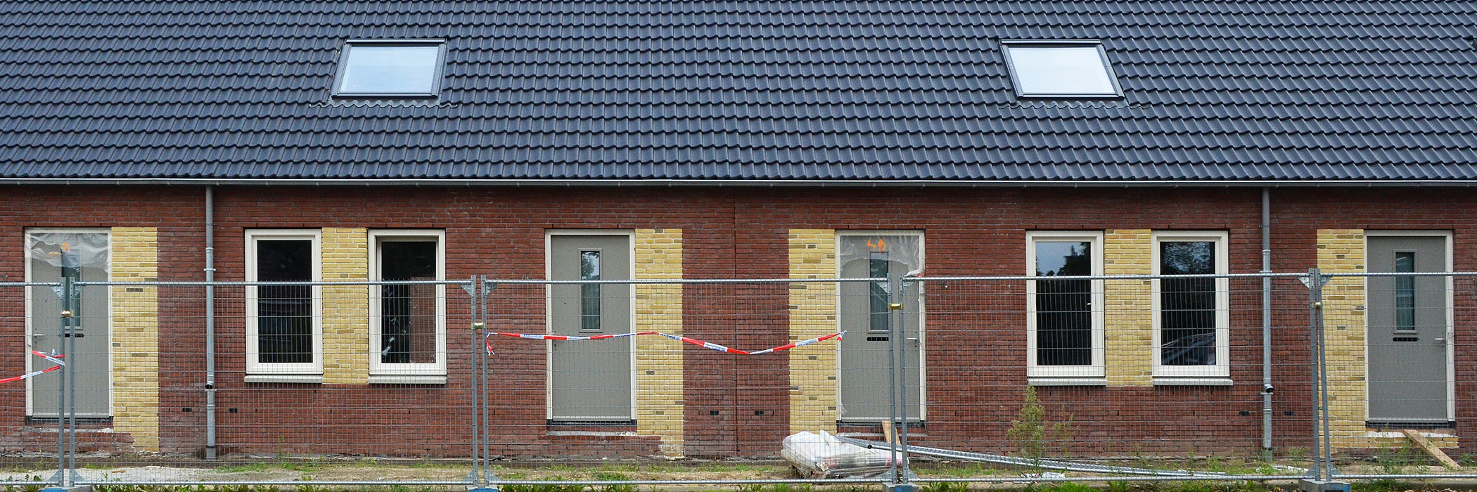 Woningen met donkerbruine bakstenen gevel, witte kozijnen, grijze deuren, zwarte dakpannen en dakramen met daarvoor een hek met een rood/wit lint