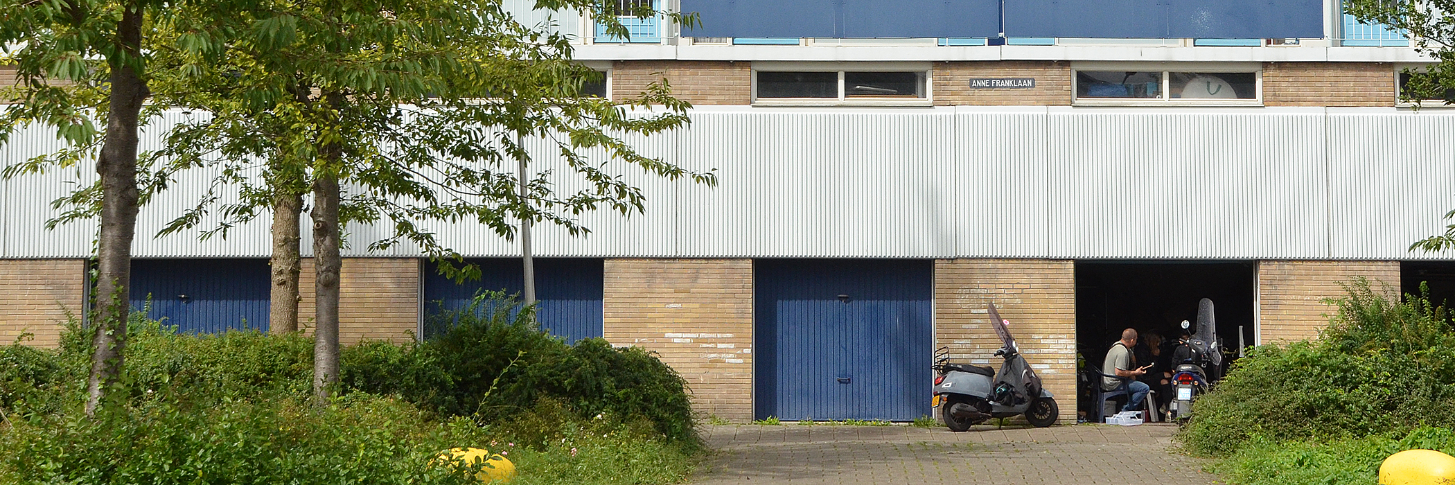 Appartementen met bruine bakstenen gevel met op de begane grond garages met blauwe overhead deuren, met twee scooters voor de deur, bosjes en bomen