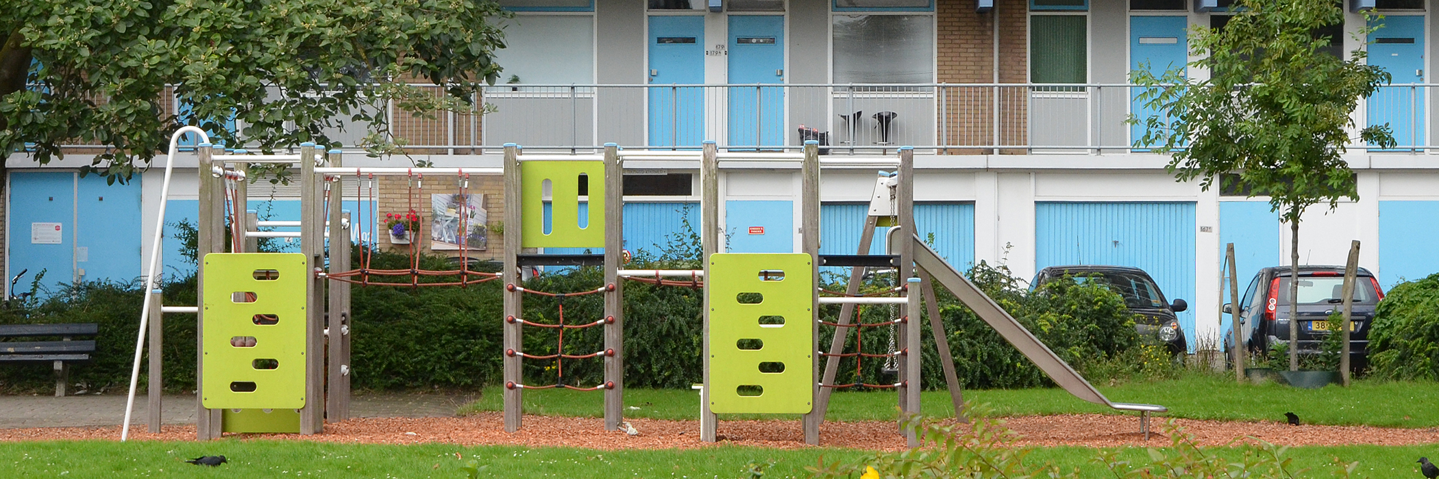 Appartementen met bruine bakstenen gevel, witte kozijnen, blauwe deuren, met op de begane grond garages met blauwe overhead deuren en een speeltuintje ervoor met een glijbaan in het gras en houtsnippers