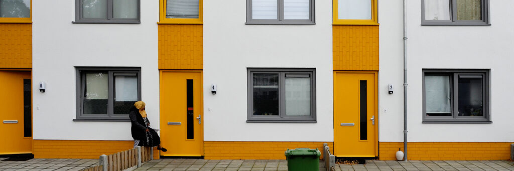 Woningen met witte gevel, gele deuren en grijze kozijnen met een vrouw die over een hekje stapt