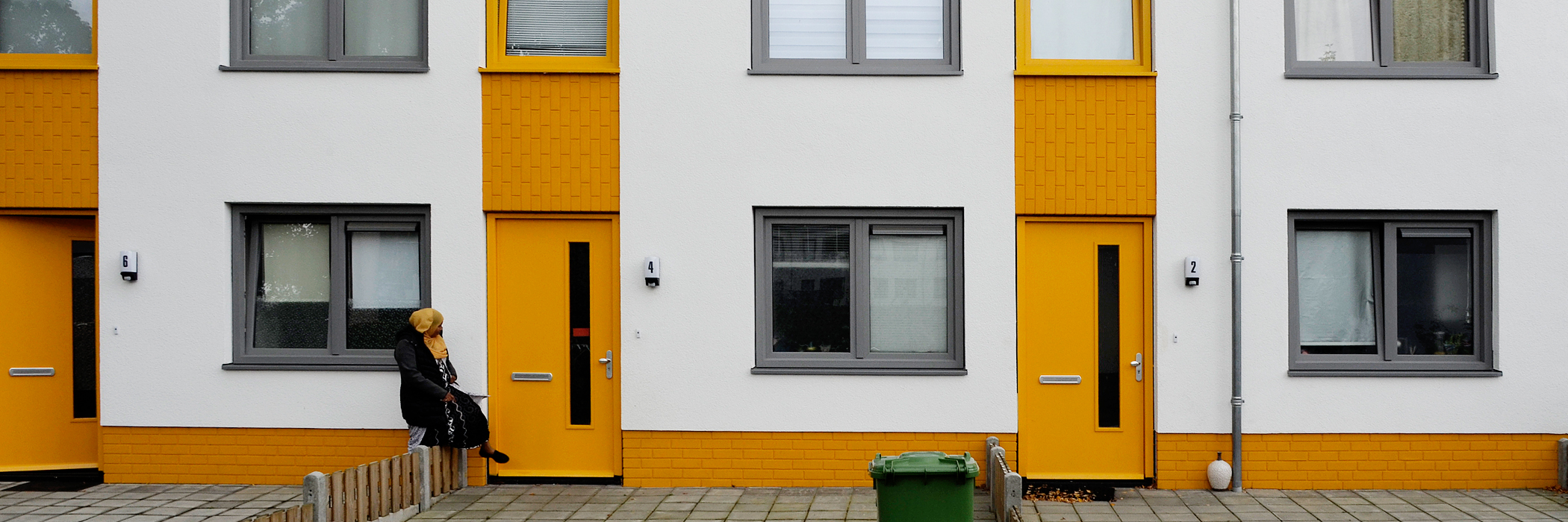 Woningen met witte gevel, gele deuren en grijze kozijnen met een vrouw die over een hekje stapt