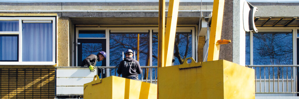 Appartementen met witte kozijnen, grijze balustrades met een gele hoogwerker ervoor waar twee mannen bij staan die aan het werken zijn