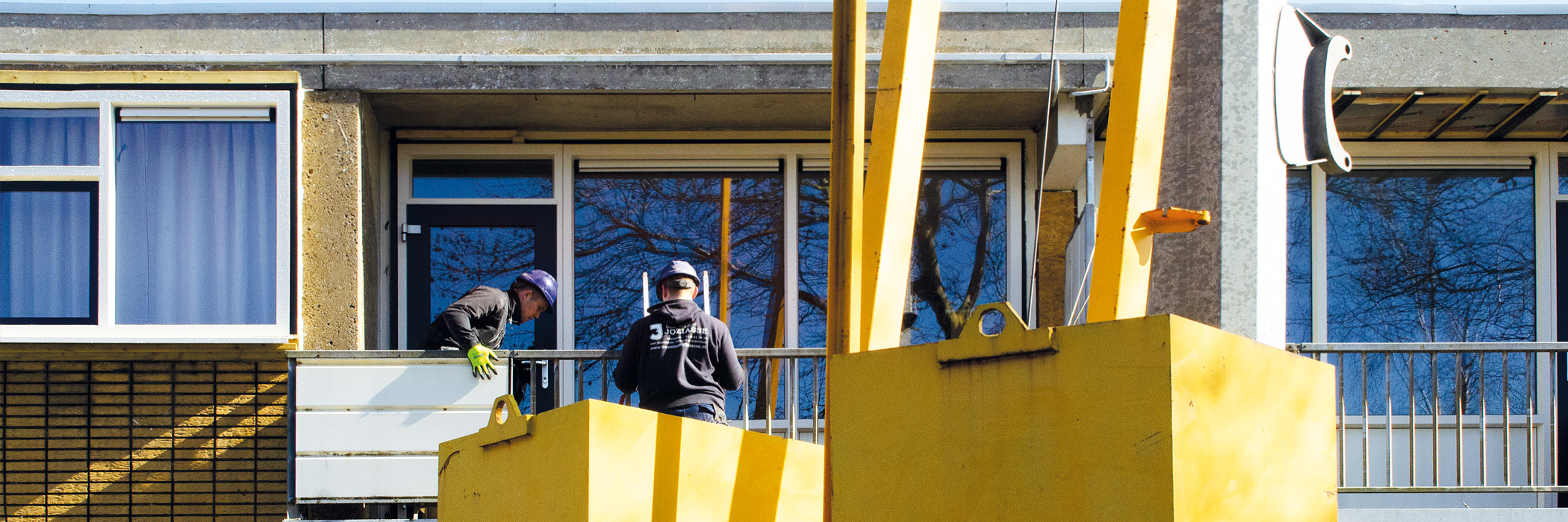Appartementen met witte kozijnen, grijze balustrades met een gele hoogwerker ervoor waar twee mannen bij staan die aan het werken zijn