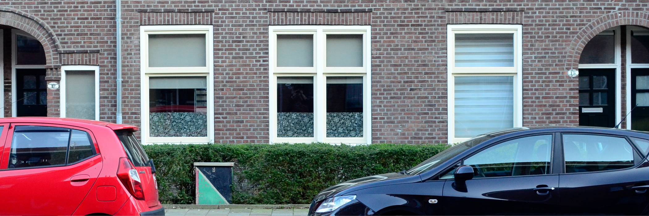 Woningen met bruin/grijze bakstenen gevel, wit/gele kozijnen, bosjes voor de deur en een grote en zwarte auto