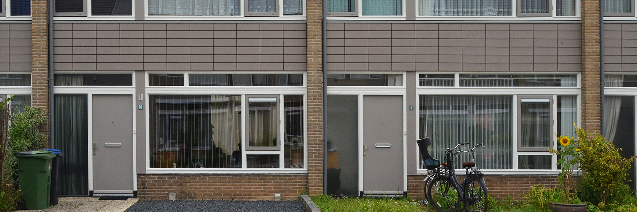 Woningen van bakstenen met grijze voordeur en een voortuin. Het stuk boven de voordeur is betegeld met grijze tegels.