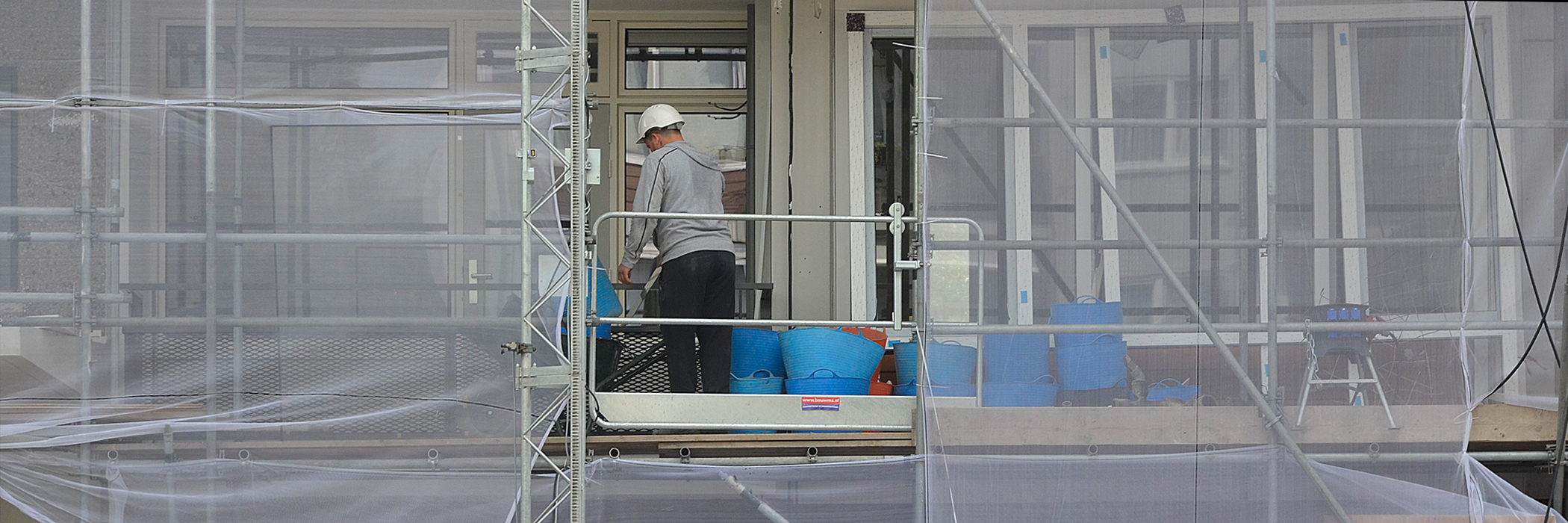 Appartementen met steigers ervoor met licht grijze doeken, waar een man op staat met een witte helm en grijs shirt en zwarte broek