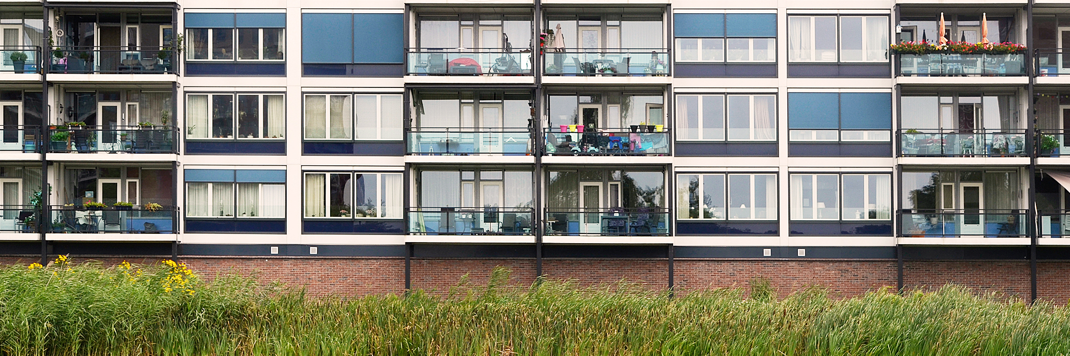 Appartementen met bruine bakstenen gevel, kozijnen in de kleuren wit en donkerblauw met blauwe zonwering en bosjes voor de deur