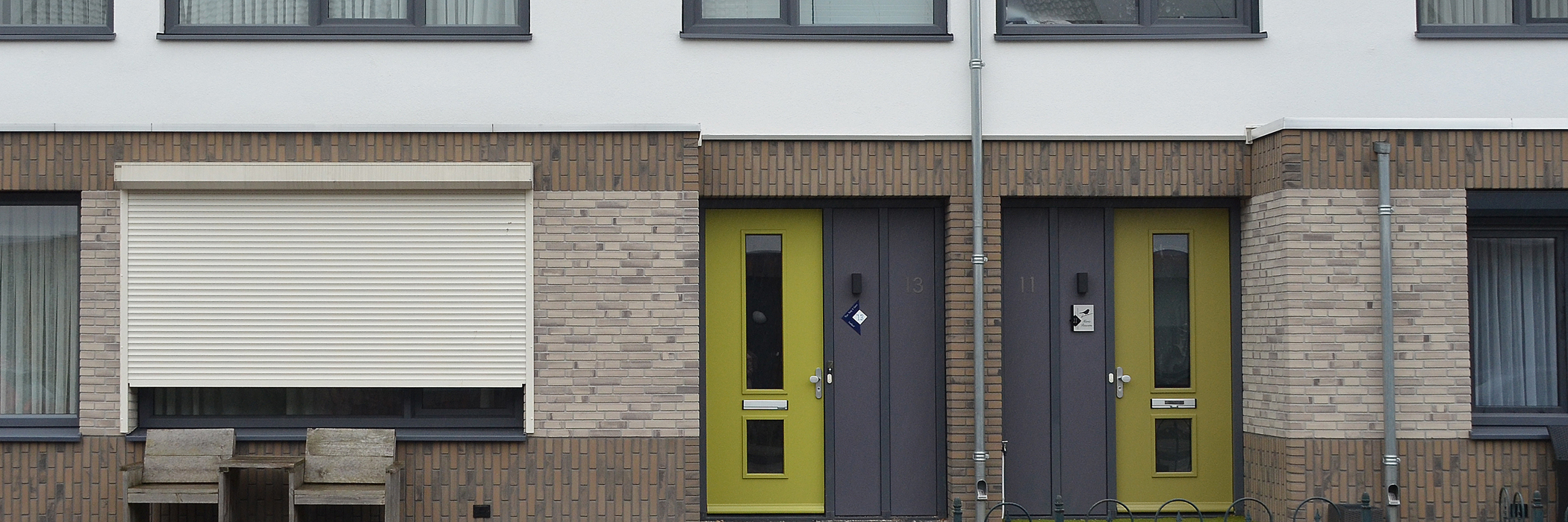 Woningen van bakstenen met een gele voordeur, witte rolluiken en donkere kozijnen.