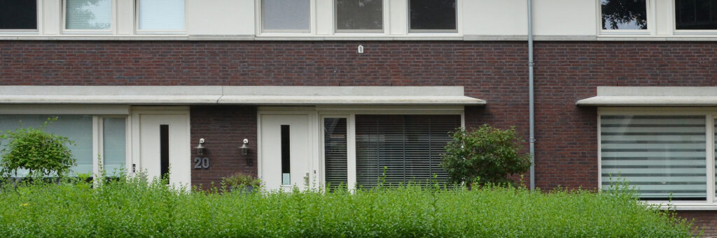 Woningen van bakstenen met een witte voordeur, witte kozijnen een bosje voor het huis.
