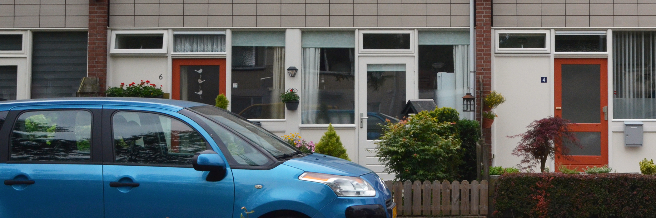 Woningen met bruine bakstenen gevel, witte kozijnen met oranje en witte deuren met een blauwe auto voor de deur