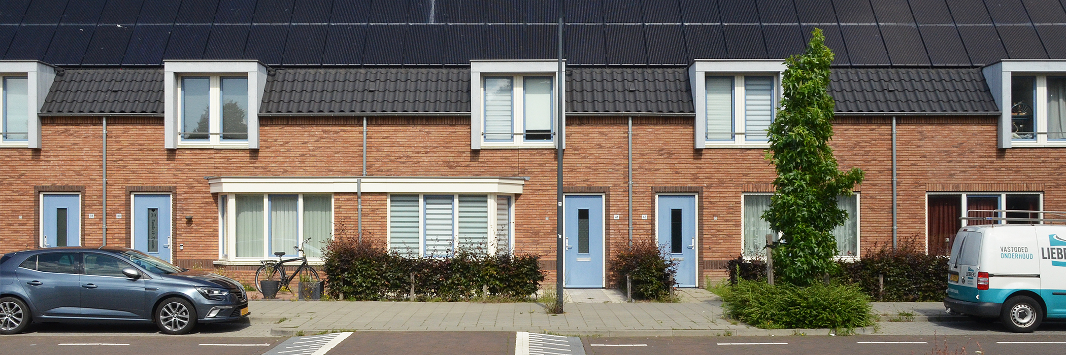 Woningen met bruine bakstenen gevel, witte kozijnen, blauwe deuren, zwarte dakpannen met zonnepanelen erop met twee auto's voor de deur