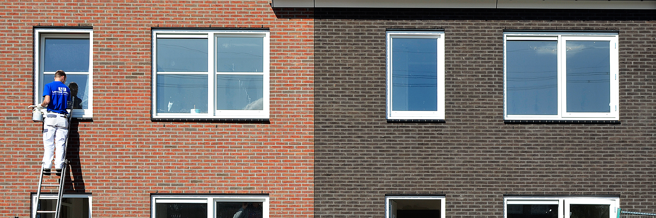 Woningen met rode en donkerbruine bakstenen gevel met witte kozijnen waar een schilder voor staat op een lader met een witte broek en zwart shirt aan