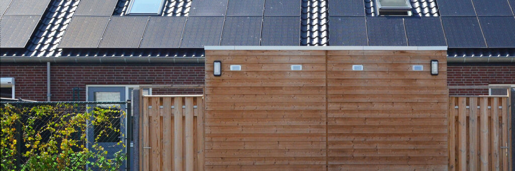 Woningen met donkerbruine bakstenen gevel, zwarte dakpannen met zonnepanelen en dakramen er op, houten schuurtjes en schuttingen