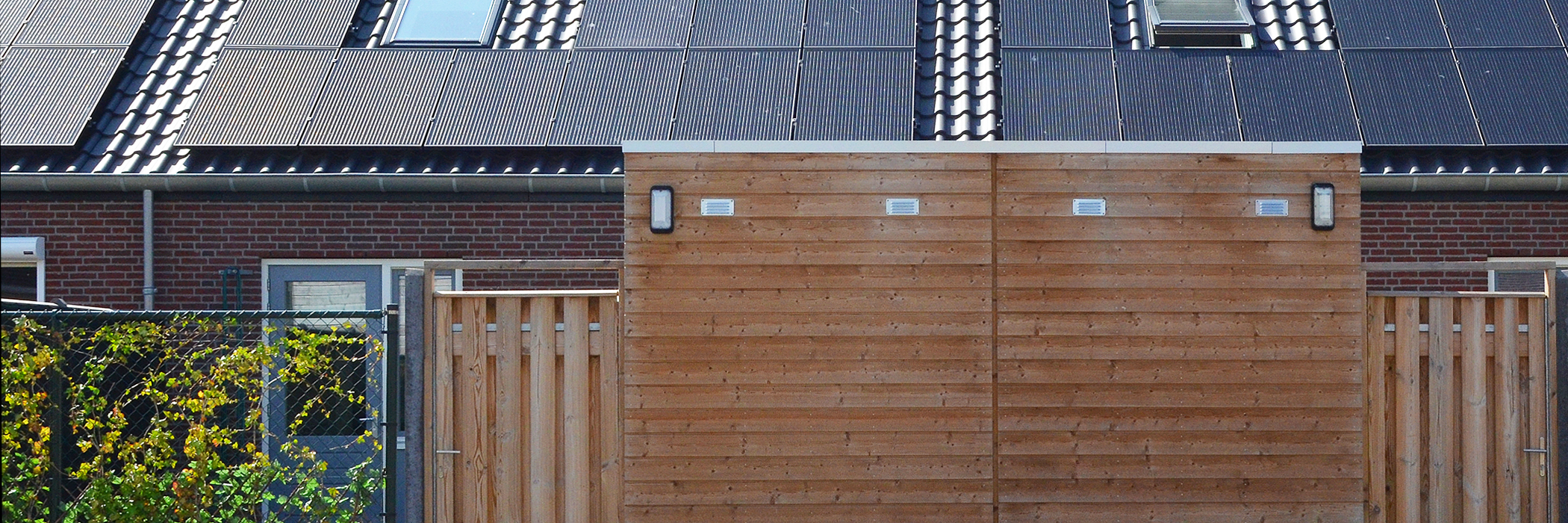 Woningen met donkerbruine bakstenen gevel, zwarte dakpannen met zonnepanelen en dakramen er op, houten schuurtjes en schuttingen