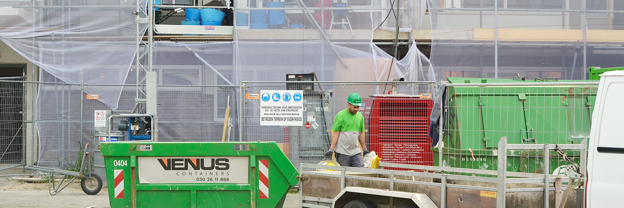 Woningen in de steigers, waar een werklui man voor staat met een groene helm, groene container en een aanhanger
