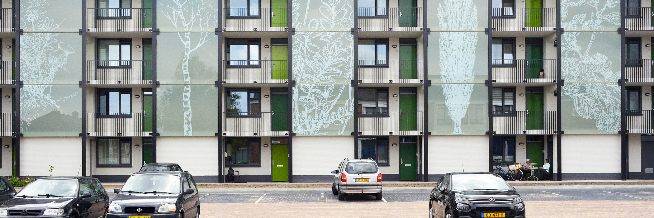 Appartementen met witte gevel, met getekend glas waar bomen op staan, donker grijze gevels, groene deuren en meerdere grijze auto's voor de deur