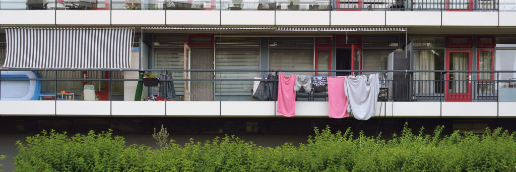 Flatgebouw met grijze kozijnen, de deuren zijn rood. aan de relingen van de balkons hangt was te drogen.