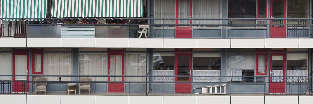 Flatgebouw met grijze kozijnen en rode deuren. Op het balkon staan stoelen.