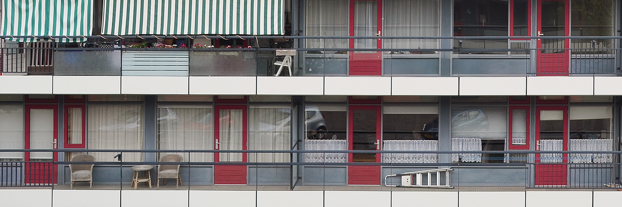 Flatgebouw met grijze kozijnen en rode deuren. op het balkon staan stoelen.