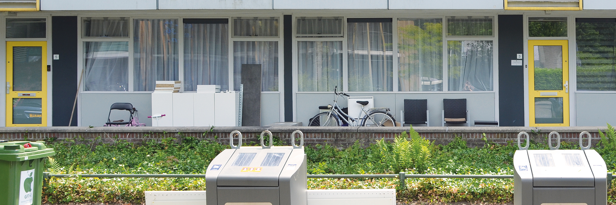 Appartementencomplex met grijze kozijnen en gele deuren, voor het complex staan afvalcontainers.