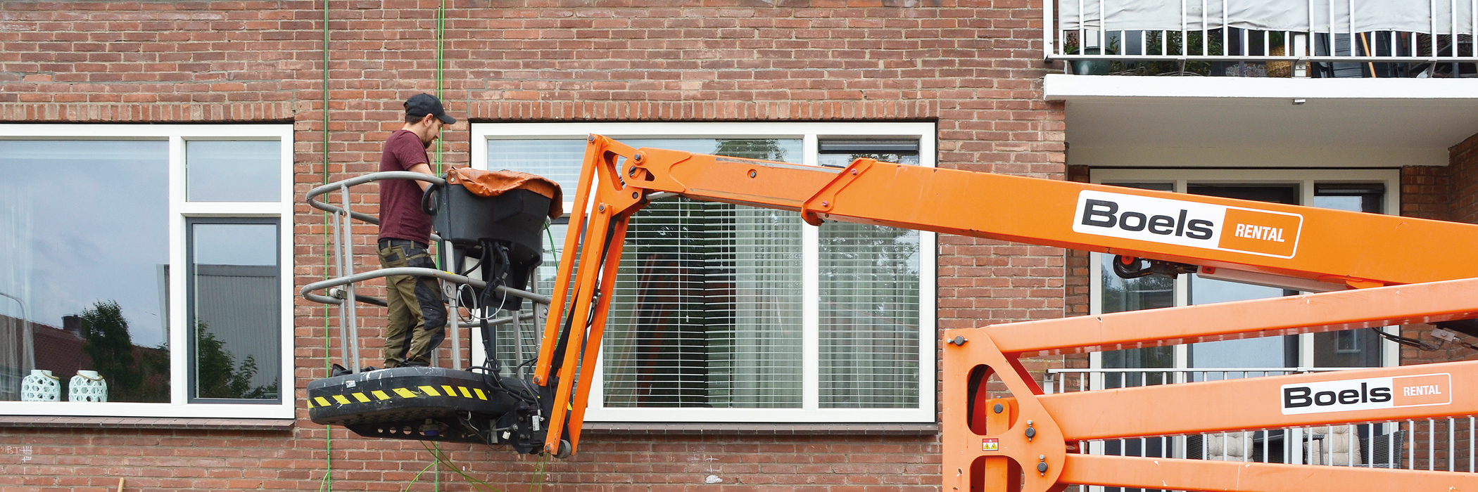 Een appartementencomplex waar een oranje hoogwerker staat met iemand in de bak die aan het werk is.